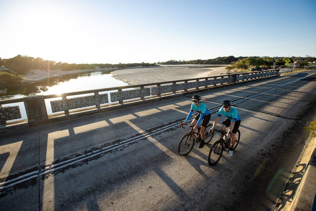 Man and woman cycling over a bridge with the sun in the background, showcasing the SYKL PRO cycling jerseys